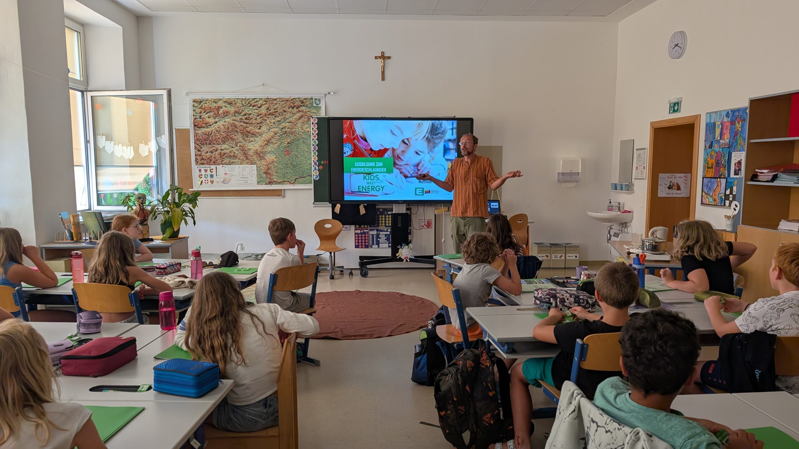 Ein Mann steht in einem Klassenzimmer vor einem großen Bildschirm. Kinder im Volksschulalter sitzen an ihren Schultischen.