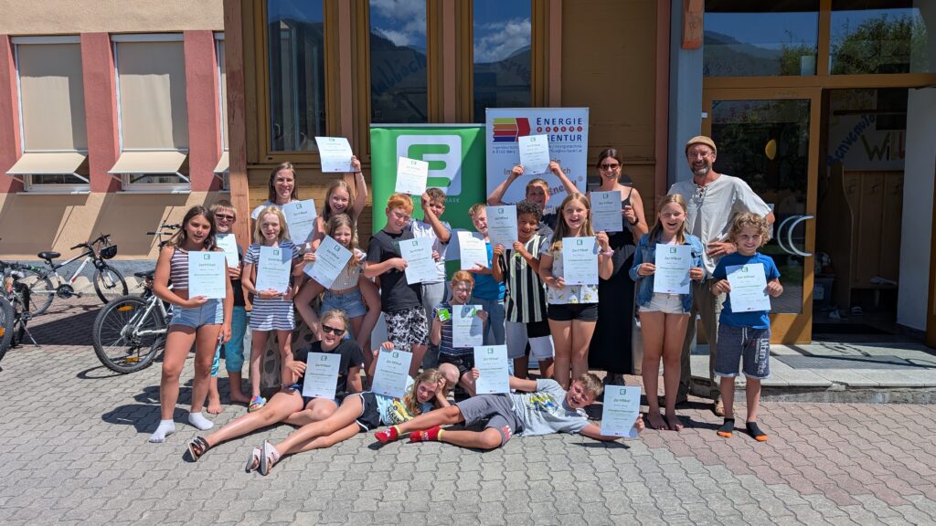 Eine Gruppe von Volksschulkindern und zwei Erwachsenen mit Urkunden in den Händen steht vor dem Schuleingang.